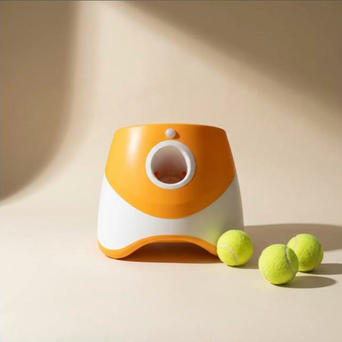 The orange and white automatic tennis ball launcher on a bone coloured surface with three tennis balls beside it.